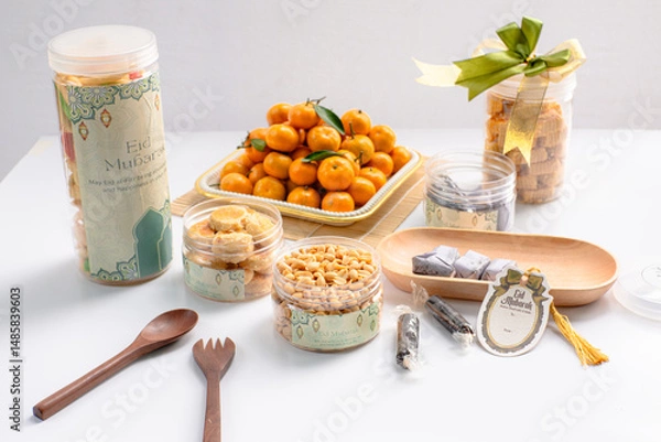 Fototapeta Eid hampers containing dry cakes in jars such as nastar, roasted peanuts without skin, oranges, and sweets on a white background with typical Eid ornaments