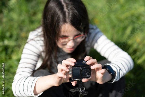 Obraz Beautiful girl squatting on a green field taking a photo on a camera. Concept of children's leisure and creativity