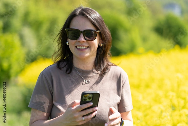 Obraz Beautiful dark-haired girl in sunglasses and a T-shirt holding a smartphone in her hands taking photos standing in front of a yellow blooming rapeseed field. Concept of digital life and nature