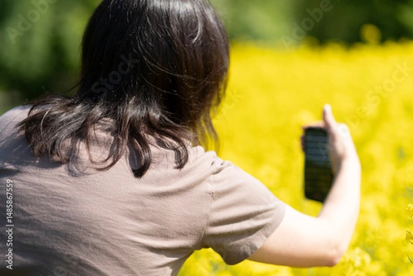 Obraz Beautiful dark-haired girl in sunglasses and a T-shirt holding a smartphone in her hands taking photos standing in front of a yellow blooming rapeseed field. Concept of digital life and nature