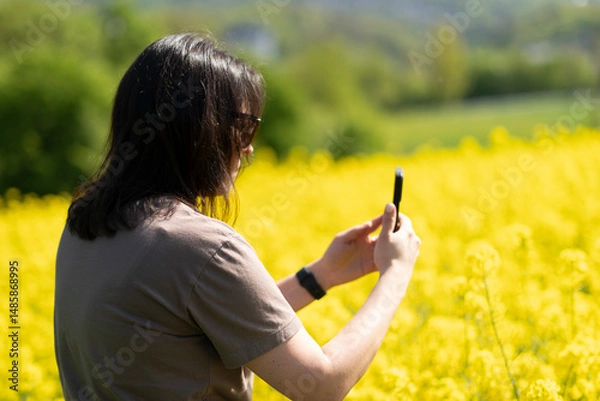 Obraz Beautiful dark-haired girl in sunglasses and a T-shirt holding a smartphone in her hands taking photos standing in front of a yellow blooming rapeseed field. Concept of digital life and nature
