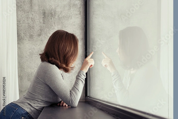 Fototapeta Portrait of beautiful сaucasian young woman touches her reflection of big panoramic window of apartments and listening music.