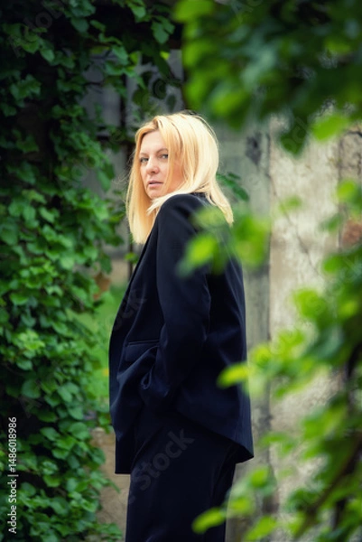 Fototapeta Stylish blonde woman in a black suit standing outdoors near ivy-covered wall. Natural greenery, elegant fashion, and serene expression in a calm urban garden scene.