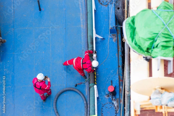 Fototapeta Top-down view of a bunker barge moored alongside a large merchant vessel. Crew in red coveralls and white helmets are busy on deck, captured during routine fuel transfer operations.