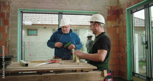 Fototapeta Construction workers examining and aligning wood piece on construction site, both wearing helmets, teamwork in building structure, focus on precision and safety