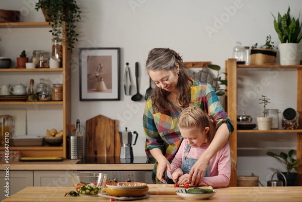 Fototapeta Mother and daughter preparing meal in cozy kitchen with wooden shelves and green plants. Mother embracing daughter while assisting in slicing vegetables on cutting board