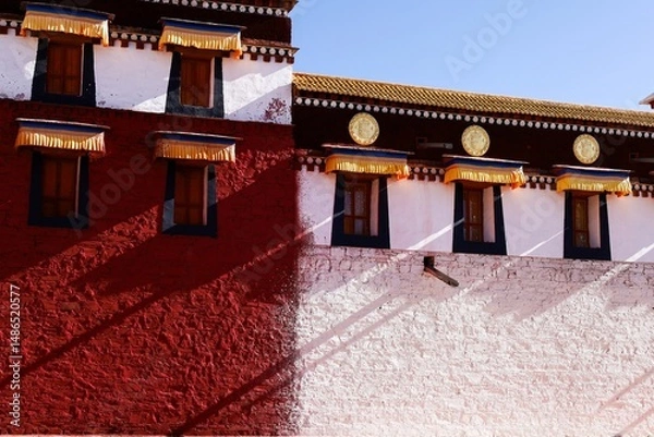 Fototapeta Traditional Tibetan architecture of Gomang Monastery in Amdo, featuring vivid red and white walls, ornate window awnings, and golden medallions, bathed in striking natural light.