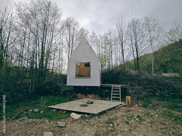 Fototapeta The white wooden board hut with big window between trees,