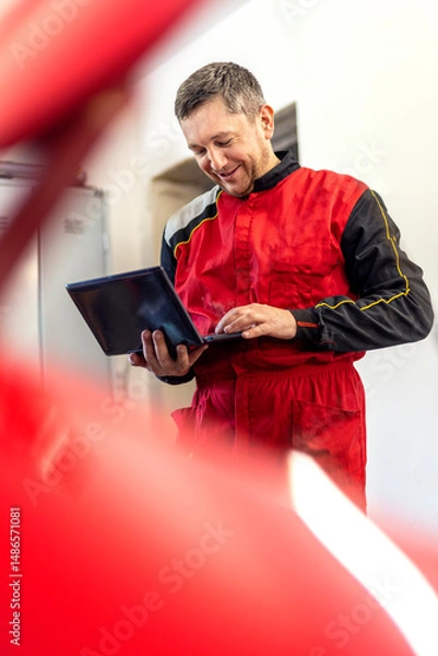 Fototapeta Car mechanic performing engine adjustment and repair using laptop. Man working with modern technology during car maintenance. Service and inspection of vehicle