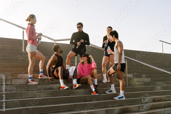 Fototapeta A multinational group of young people is taking a break while sitting and standing on stairs outdoors on a sunny day, wearing athletic clothing after a training session near a modern building.
