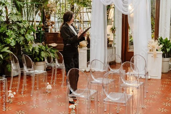 Fototapeta A woman stands in a beautiful indoor space filled with plants, transparent chairs, and elegant decor, taking notes with a planchette. The concept of an intimate event setup.