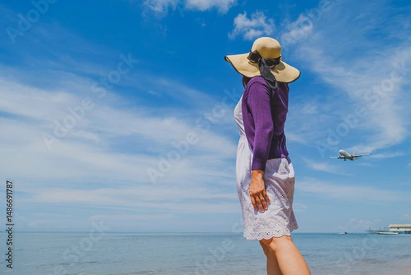Fototapeta a woman wearing a straw hat near the beach look up to the sky when the airplane passing by..