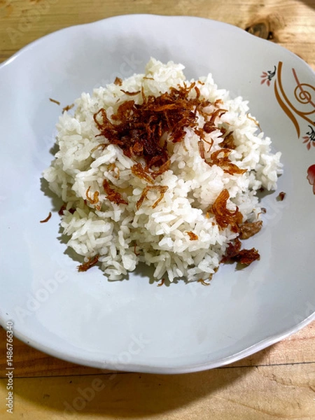 Fototapeta White rice with fried onions on top on a white plate with floral motifs on a wooden table. rice as food