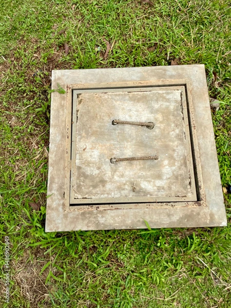 Fototapeta Grey cement control tank cover with iron handle on top in a garden with green grass around it, drainage control tank concept