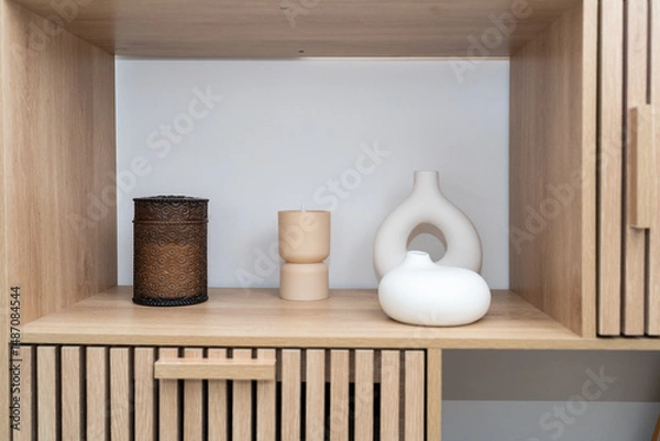 Obraz A wooden shelf holds an assortment of decorative items including a textured container, a candle holder, and a smooth ceramic piece. The arrangement is visually appealing and minimalist.