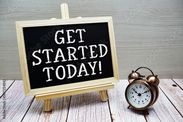 Fototapeta Get Started Today! text written on chalkboard with alarm clock on wooden background
