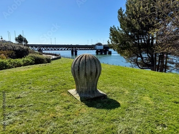 Fototapeta Stone bollard on waterfront grass with bridge backdrop