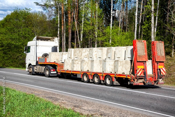 Fototapeta A white truck with a lowboy semitrailer transports concrete products along a country road against a forest background. Oversized cargo. Copy space for text