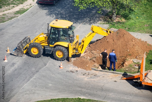 Fototapeta A yellow excavator dug up a road to lay electrical cables, workers stand near the hole, industry. Copy space for text