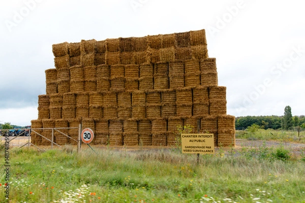 Fototapeta In a tranquil field in France, a tall structure of straw bales stands stacked against a backdrop of overcast clouds. The area is marked for public safety.