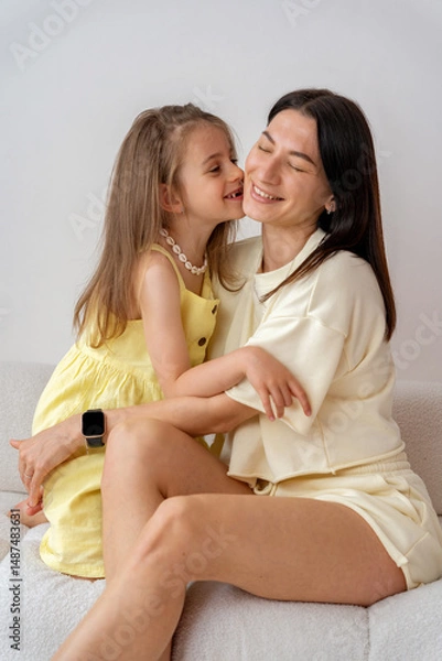 Fototapeta Happy mother and child. Daughter kissing mom on cheek and hugging at home in white room. Family time. Mother's Day.