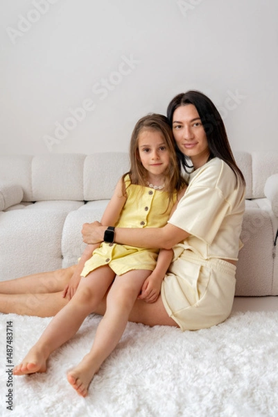 Fototapeta Happy mother hugging her daughter while sitting on white carpet at home in white interior. Mother's love. Family relationships and Mother's Day.