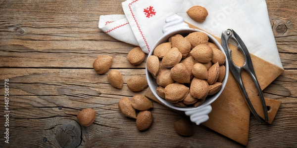 Fototapeta Whole almonds in shell in white ceramic bowl with old nutcracker on rustic wooden background, top view, space for text.