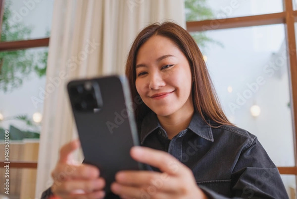 Fototapeta Portrait image of a young woman holding and using mobile phone in cafe