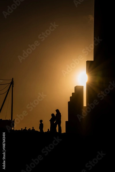 Fototapeta A silhouette of a family standing in front of a building. The sun is shining on the building, creating a warm and inviting atmosphere