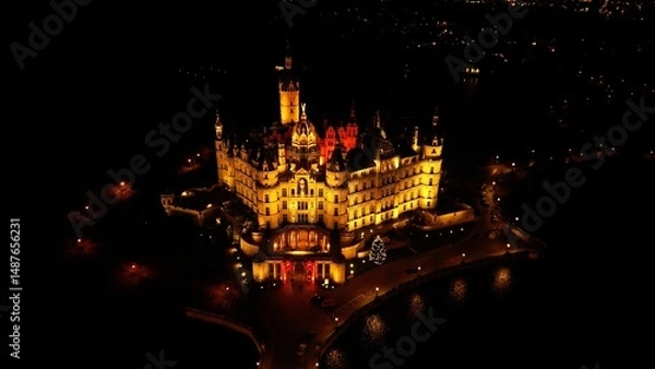 Fototapeta A night drone photo of Schwerin Castle in Northern Germany. The castle is beautifully lit and reflected in the surrounding lake. A peaceful, atmospheric image capturing historic architecture and winte
