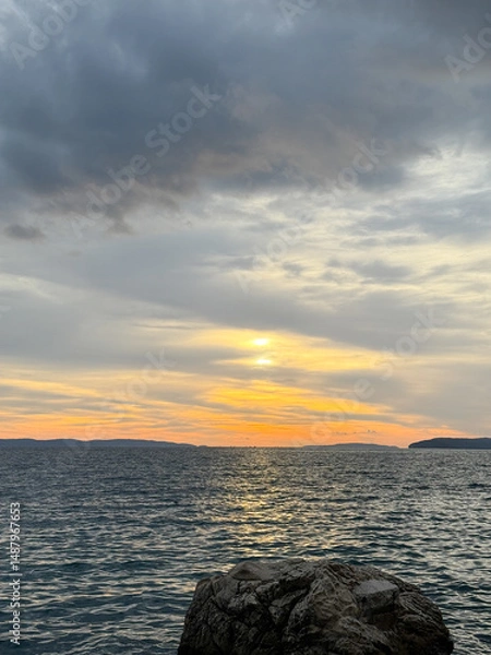 Fototapeta Golden sunset illuminating clouds over calm sea with rock in foreground