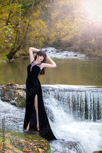 Fototapeta A brunette in a posh black dress posing by the river with a small waterfall.
