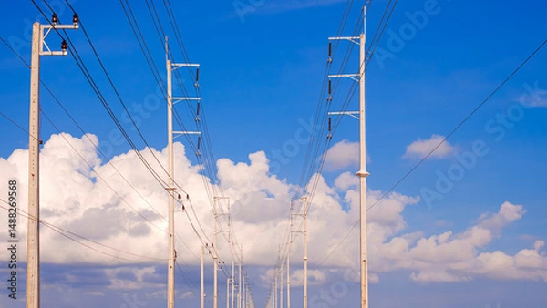 Fototapeta Two rows of electric poles with cable lines on the road against white clouds on blue sky background, low angle and front view with copy space
