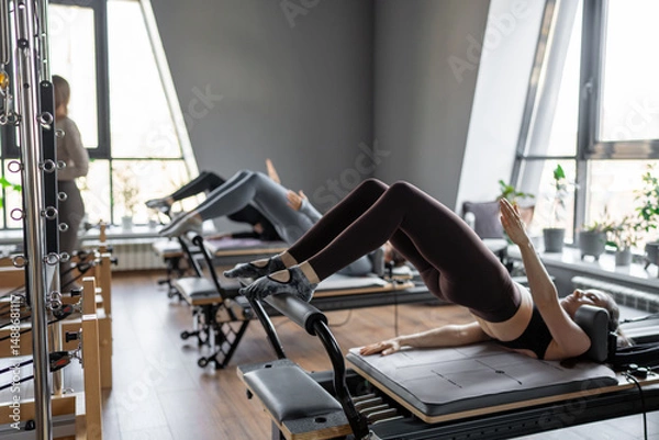 Fototapeta A group of woman exercises Glute bridge on a reformer bed machine in natural sunlight, focusing on pelvis and hips.