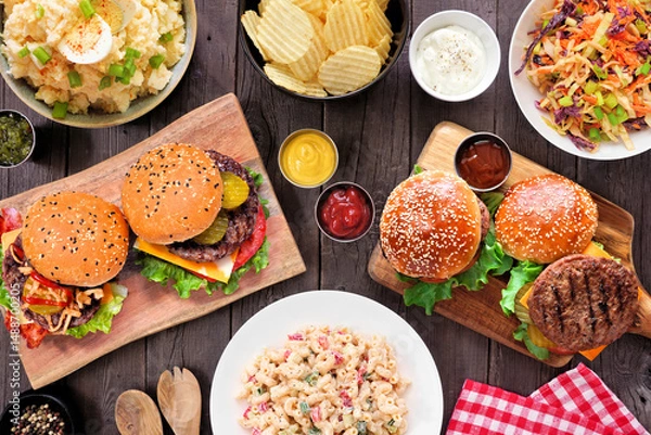 Obraz BBQ table scene with hamburgers, classic salads and snacks. Top view over a dark wood background.