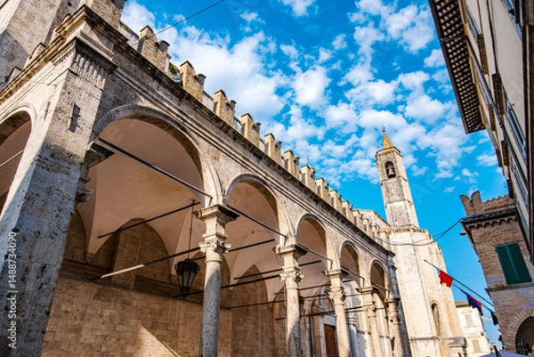 Fototapeta Loggia dei Mercanti featuring travertine arches and columns, framed by the bell tower of the Church of San Francesco, creates a picturesque scene under a cloudy blue sky