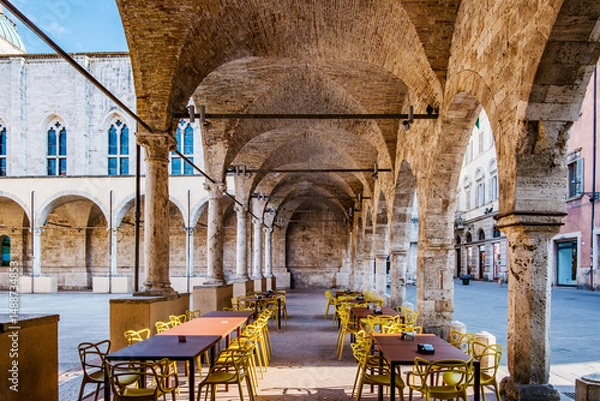 Fototapeta Empty tables and yellow chairs lined under brick archways in the loggia of Piazza San Francesco, Ascoli Piceno, create a serene urban architectural scene