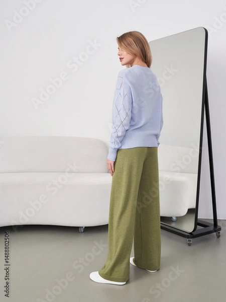 Fototapeta A woman with light brown hair in a blue sweater and olive-green pants stands sideways. She looks at a full-length mirror, while a stylish sofa is visible in the background