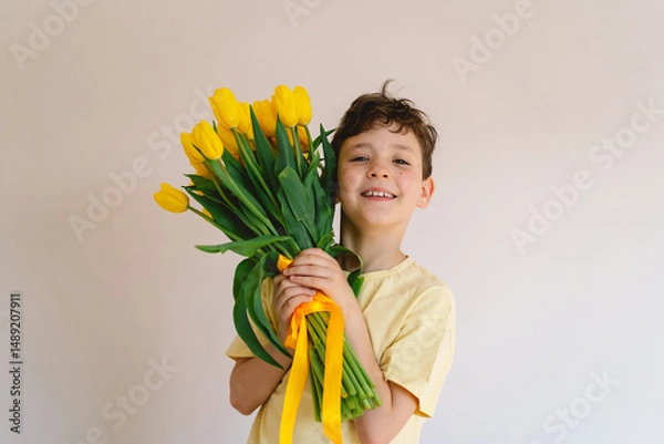 Obraz A boy stands indoors holding a large bouquet of yellow tulips, radiating happiness with a big smile on his face. The bright atmosphere reflects cheerfulness and warmth. Mother's Day and spring concept