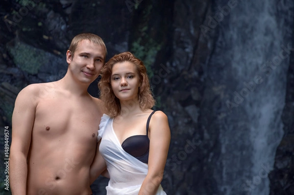 Fototapeta A young semi-naked couple looks at the camera, standing by a waterfall in a grotto. Close-up. Copy space