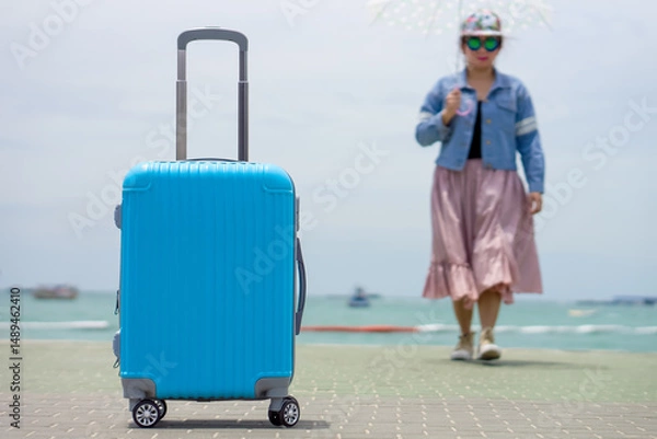 Fototapeta Bright Blue Luggage on a Pier with a Traveler and Sea View. Vibrant blue hard-shell suitcase stands prominently on a seaside pier, with a person holding an umbrella walking in the blurred background