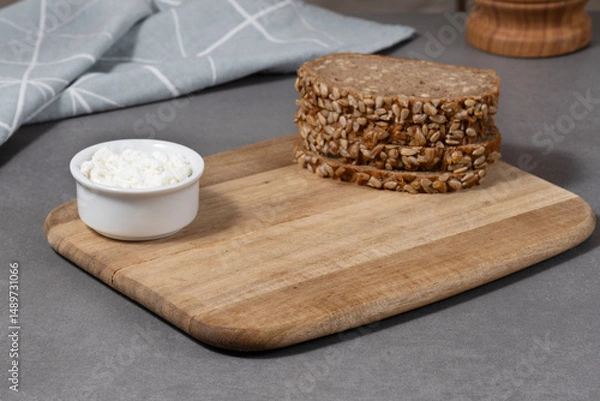 Fototapeta Slices of whole grain bread with sunflower seeds and a small bowl of cottage cheese on a wooden cutting board