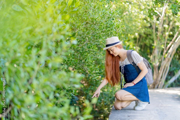 Fototapeta Young woman in hat and backpack kneeling and pointing at nature along green forest path, exploring flora during a peaceful summer outdoor adventure.
