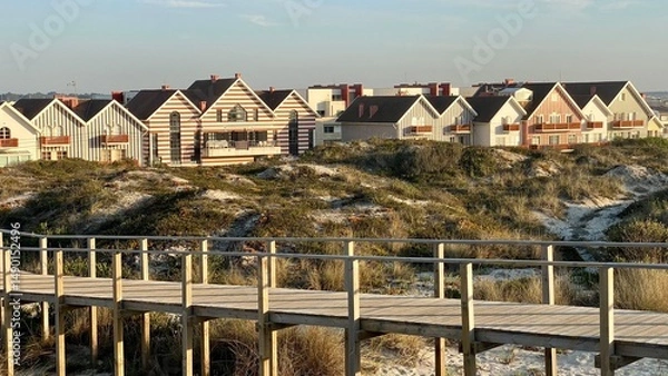 Fototapeta Colorful beach houses along a wooden path in Costa Nova, Portugal — charming seaside village with iconic striped buildings and ocean breeze