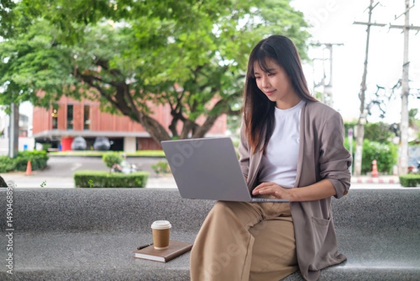 Fototapeta Woman in smart casual outfit using computer at a park bench, representing freelance or digital nomad lifestyle.
