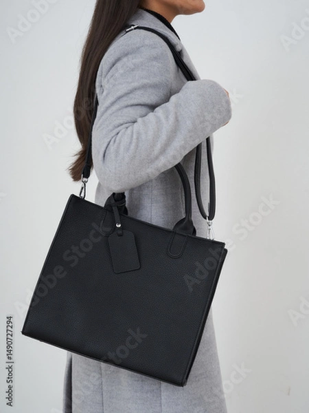 Fototapeta A person in a light gray coat carries a sleek black leather handbag with a shoulder strap and a blank tag. The hair is long and dark, contrasting with the outfit and accessory