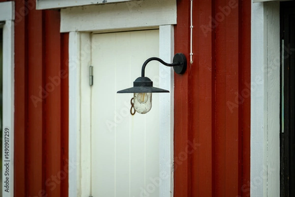 Obraz A black outdoor light hangs beside a white door on a red wooden wall. The rustic atmosphere highlights the charm of the structure in a serene environment