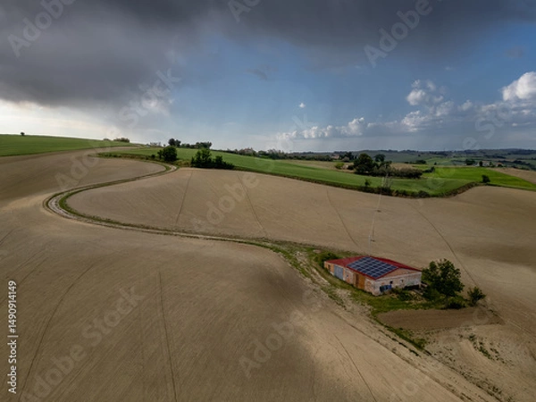 Fototapeta Italy, May 15, 2025: Aerial view of a farmhouse equipped with solar panels in the middle of plowed fields. Peace, tranquility with renewable energy is conveyed by this image
