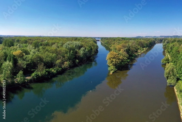 Fototapeta Die Lechmündung in die Donau zeigt eine friedliche Flusslandschaft mit üppiger Vegetation und klarem Wasser. Die Spiegelung der Bäume im Wasser und der blaue Himmel schaffen eine idyllische Atmosphäre