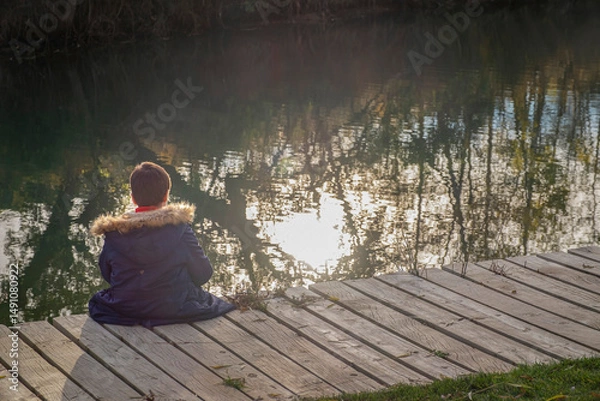 Obraz A child in a winter coat sits alone on a wooden dock, looking at the calm water reflecting trees and sunlight, creating a peaceful and contemplative mood